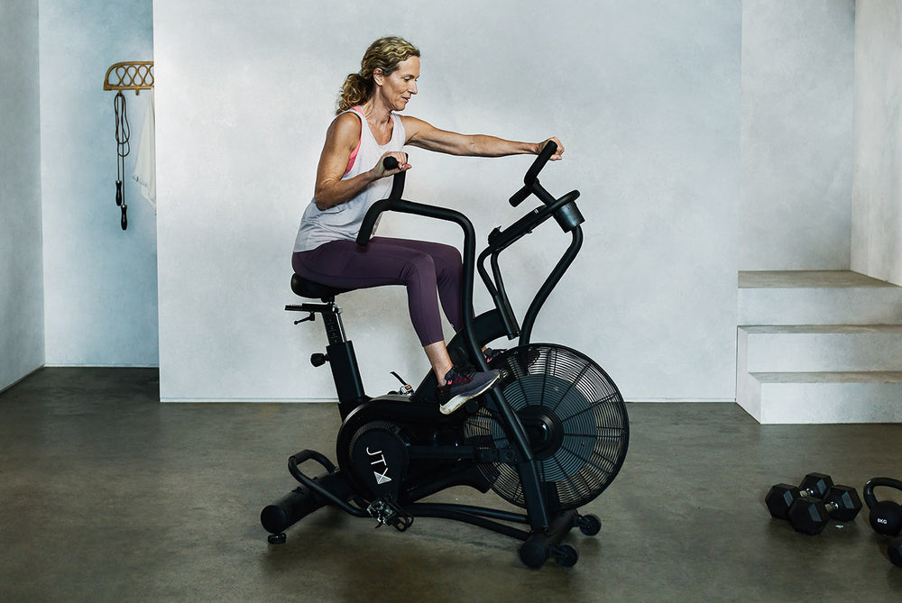 Woman exercising on a JTX Mission Air bike in a home gym. Her feet are resting on the foot pegs while she moves the handlebars.