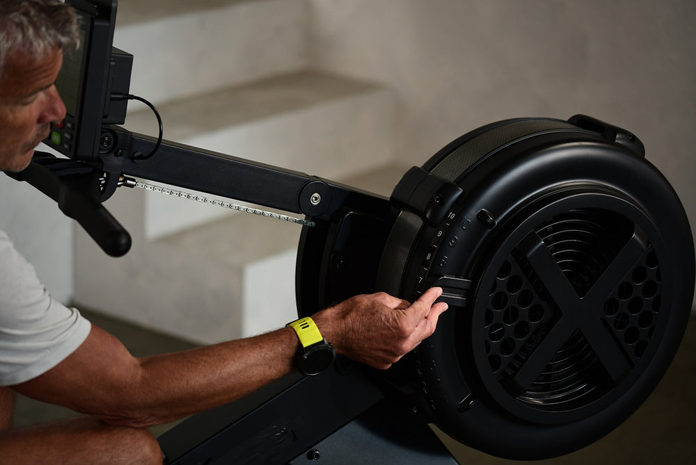 Man manually adjusting the resistance setting on the front fan wheel of a JTX Ignite AirX Rower.