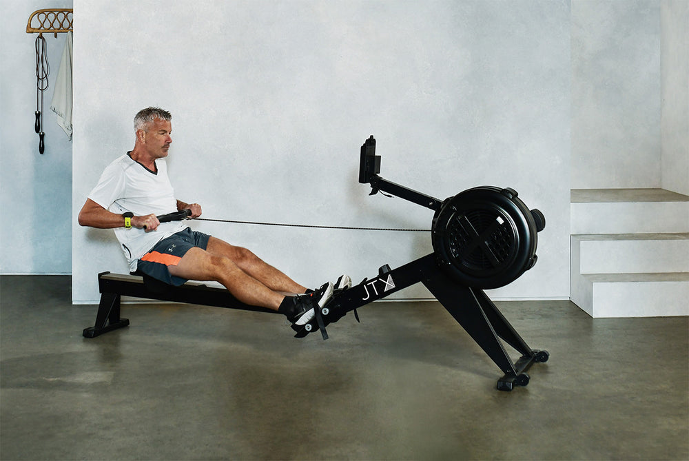 Man using a JTX Ignite AirX Rower air rowing machine in a home gym setting. His legs are fully extended as he performs a pulling motion.