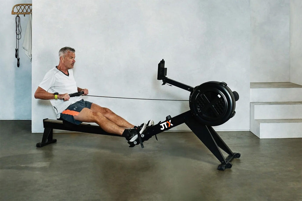 Man using a JTX Ignite AirX Rower air rowing machine in a home gym setting. His legs are fully extended as he performs a pulling motion.