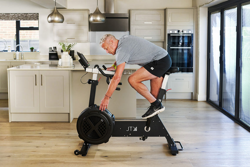 Man sat on a JTX Ignite AirX Rider fan bike while leaning forward to adjust the resistance setting on the front mounted fan wheel in his kitchen.