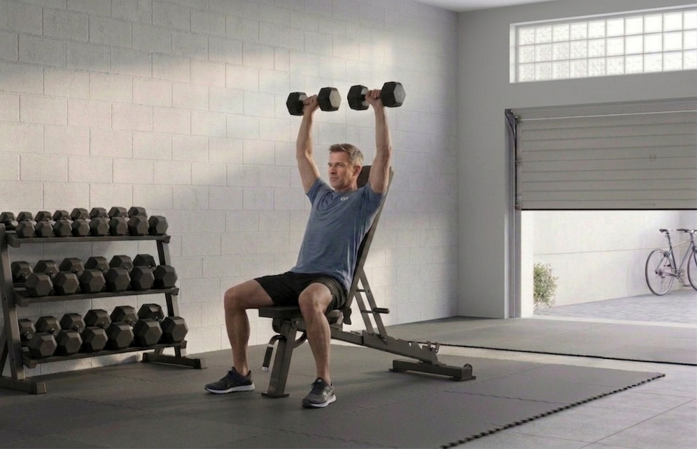 Man using the JTX FID bench to complete a dumbbell shoulder press exercise in a garage home gym