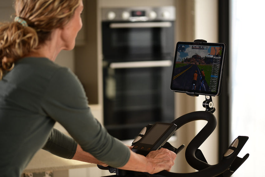 Woman using a JTX Studio Pro exercise bike at home while following a virtual Zwift cycling route on a tablet.