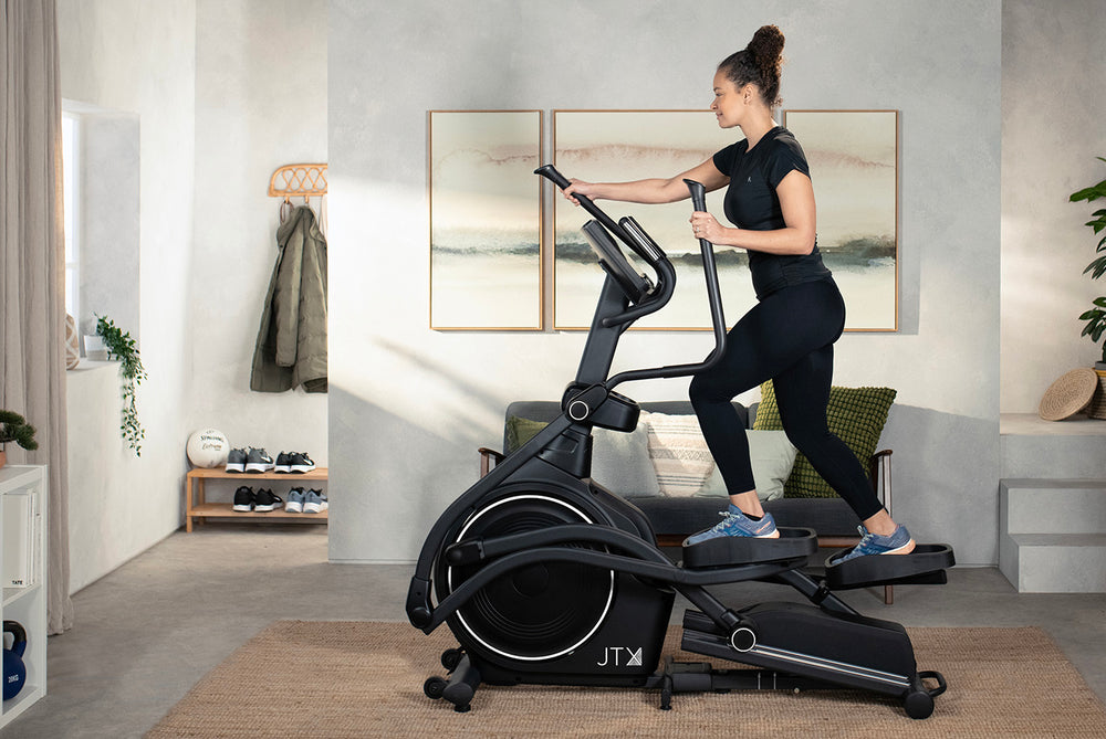 Woman exercising on a JTX Zenith Rise cross trainer in a modern living room.