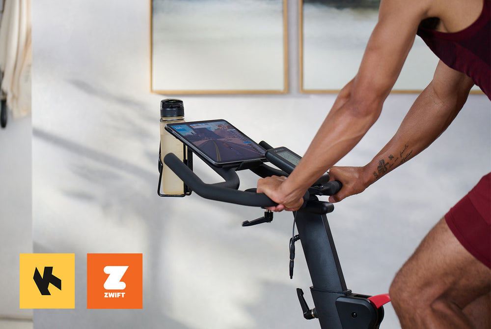 Man on an exercise bike following a Zwift route on their tablet. Kinomap and Zwift logos are on display.