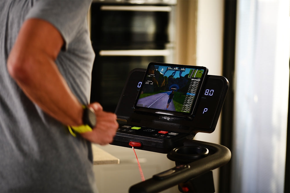 Man jogging on a JTX RunRise treadmill at home while connected to the Zwift app on a tablet.