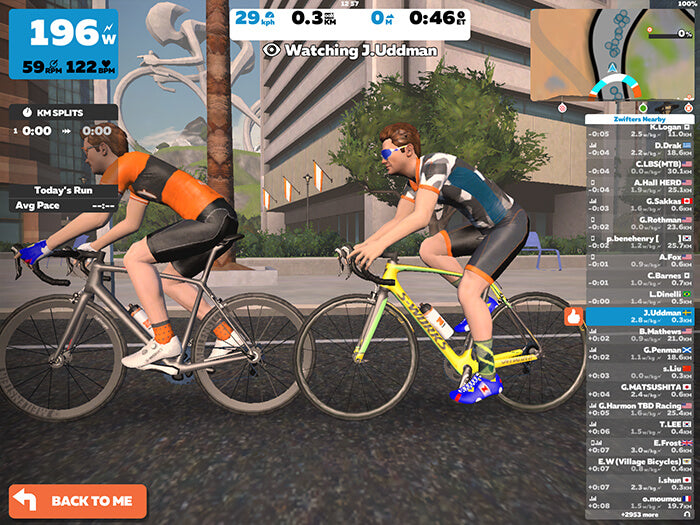 Zwift virtual cycling workout showing two riders racing through a digital city environment with live performance metrics and leaderboard data on screen.