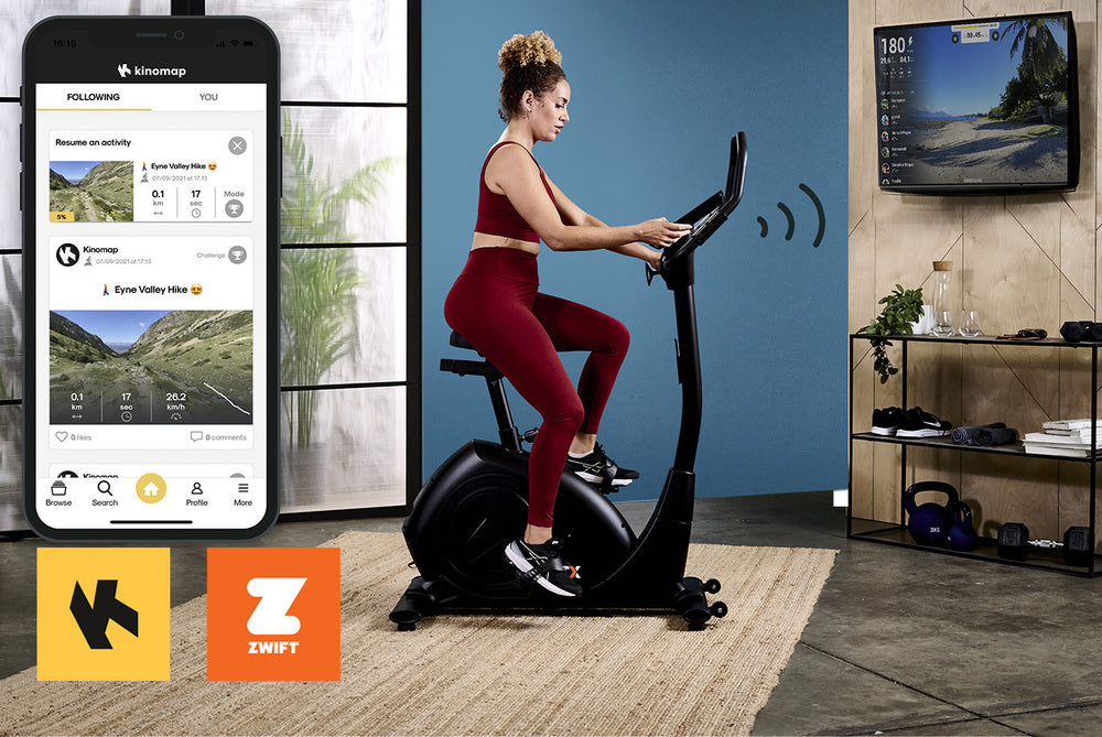 Woman exercising on a JTX Cyclo-Go  X upright bike. A smart phone is displaying the Kinomap app menu. The Kinomap and Zwift logo are displayed in the bottom left corner of the image.