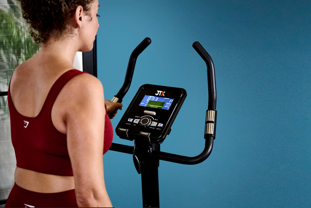 Computer console and handlebars of the JTX Cyclo-Go X upright bike while a woman is using it.