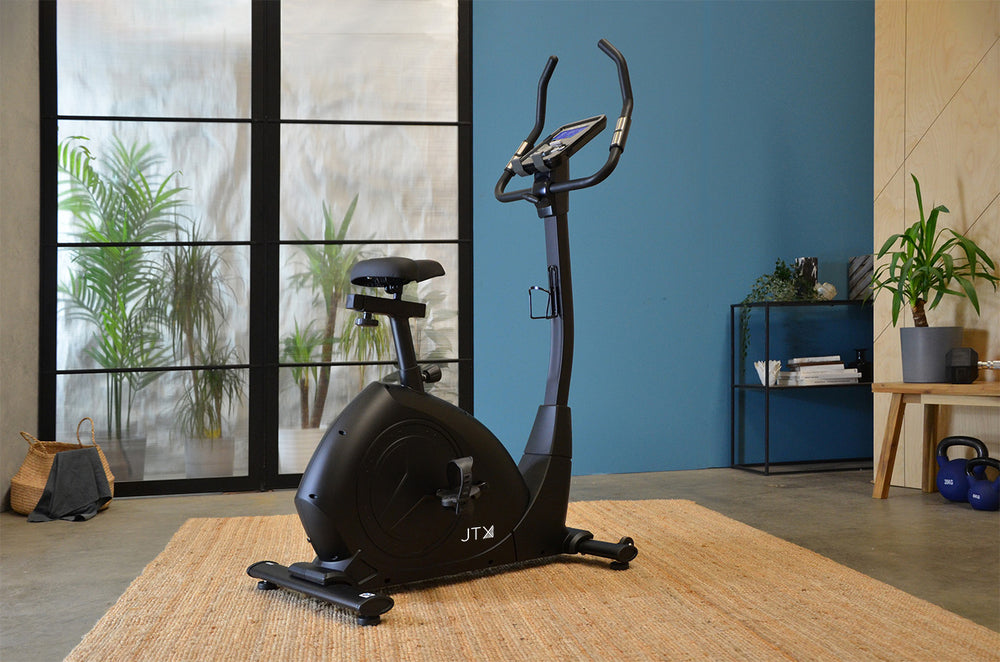 JTX Cyclo-Go X upright bike in a home setting with a blue wall and large windows.