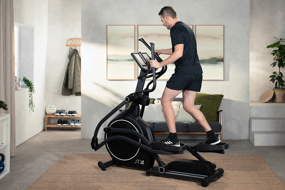 Man exercising on a JTX Zenith Rise cross trainer in a modern living room.